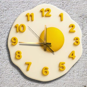 Pode incluir: Um relógio de parede extravagante em forma de ovo frito. O relógio apresenta uma gema amarela, uma clara de ovo branca e algarismos amarelos em relevo. Os ponteiros do relógio são dourados. O relógio está montado numa parede cinzenta texturizada.