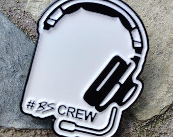May include: A white and black enamel pin featuring a headset design. The pin has a white background with black outlines and details, including the headset and a microphone. The text "#BS CREW" is displayed on the pin.