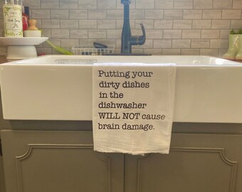 May include: A white rectangular kitchen towel draped over a white farmhouse sink. The towel features black printed text that reads: Putting your dirty dishes in the dishwasher WILL NOT cause brain damage. The fabric has a smooth, clean appearance.