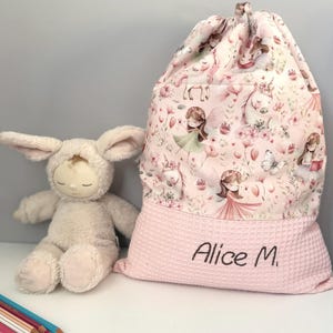 Può includere: Una borsa rosa con coulisse con un motivo floreale e di unicorni, e il nome "Alice M." ricamato sul fondo. Un coniglietto di peluche e matite colorate sono accanto alla borsa.