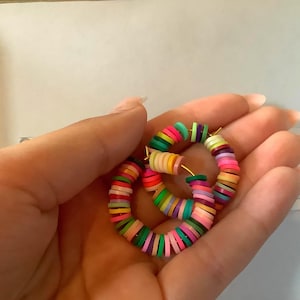 Puede incluir: Un par de pendientes de aro coloridos. Los pendientes están hechos de pequeñas cuentas redondas y planas de varios colores, incluyendo rosa, verde, amarillo y morado. Las cuentas están ensartadas para formar una forma circular, con aros de metal dorado.