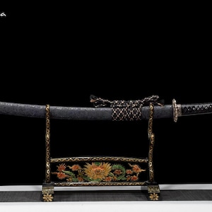 May include: A black Japanese katana sword with a textured blade, resting on a decorative stand. The stand is black with gold accents and a colorful dragon design. The sword is tied with a decorative cord.