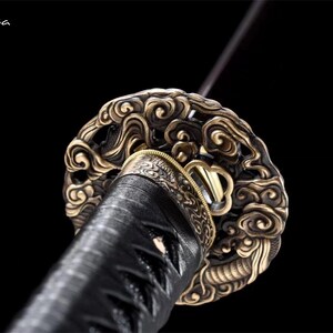 May include: Close-up of a katana sword hilt. The hilt features an ornate, swirling design in gold and bronze tones. The handle is wrapped in black material, and the blade extends upwards. The background is black.