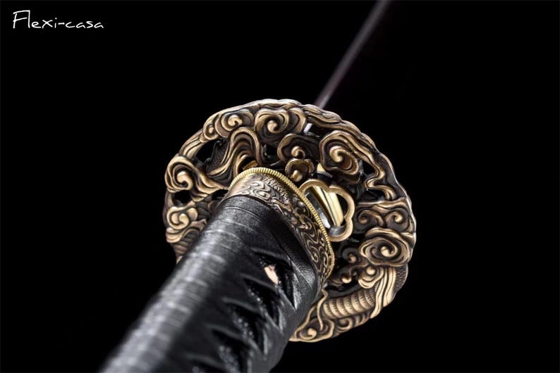 May include: Close-up of a katana sword hilt. The hilt features an ornate, swirling design in gold and bronze tones. The handle is wrapped in black material, and the blade extends upwards. The background is black.