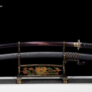 May include: A Japanese katana sword with a purple blade and black handle rests on a decorative stand. The scabbard is black with a textured pattern. The stand is black with gold accents and a floral design. The text "Flexi-casa" is in the upper left corner.