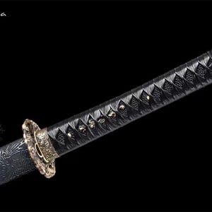 May include: Close-up of a katana sword. The handle is wrapped in black material with a diamond pattern. The guard is gold-colored with intricate designs. The blade is not visible. The sword is set against a black background.