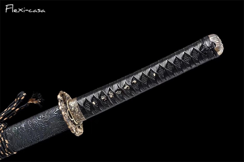 May include: Close-up of a katana sword. The handle is wrapped in black material with a diamond pattern. The guard is gold-colored with intricate designs. The blade is not visible. The sword is set against a black background.