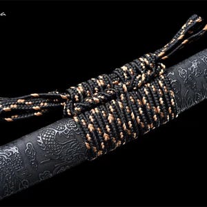 May include: Close-up of a black and gold wrapped sword handle. The handle is wrapped with a black and gold cord. The sword blade has a detailed dragon pattern. The text "Flexi-casa" is in the upper left corner.