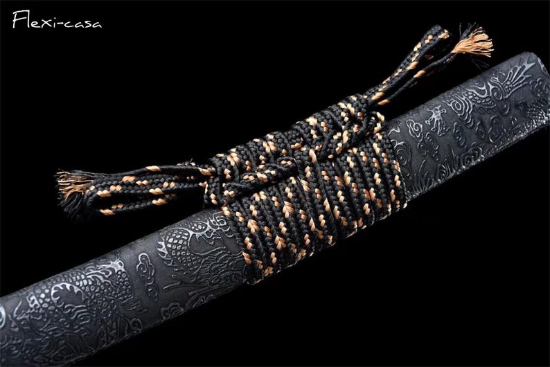 May include: Close-up of a black and gold wrapped sword handle. The handle is wrapped with a black and gold cord. The sword blade has a detailed dragon pattern. The text "Flexi-casa" is in the upper left corner.