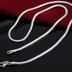 May include: A silver chain necklace with a textured, woven design. The necklace is displayed on a dark surface with a red cloth in the background. The necklace has a clasp closure.