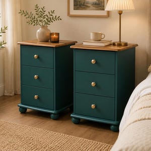 May include: Two matching bedside tables featuring a deep teal finish and a natural wood top. Each unit has three drawers equipped with small, round, gold-toned metal knobs. The tables are supported by four rounded, ball-shaped feet at the base.