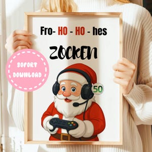 Könnte beinhalten: Ein gerahmter Druck mit einem Cartoon von Santa Claus, der ein Headset trägt und einen Gamecontroller hält. Der Druck enthält den Text "Fro- HO - HO - hes ZOCKEN" und einen rosa Kreis mit dem Text "SOFORT DOWNLOAD". Santa trägt einen roten Anzug und einen weißen Bart.