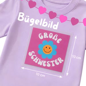 Könnte beinhalten: Ein hellviolettes T-Shirt mit einem rosa Bügelbild, das eine blaue Blume, ein gelbes Smiley-Gesicht und den Text "GROBE SCHWESTER" zeigt. Das Bügelbild misst 10 cm mal 10 cm. Über dem Motiv ist eine Kette aus rosa und magentafarbenen Herzen abgebildet.