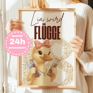 May include: A framed print featuring a cartoon duckling with a pink bow holding Euro banknotes. The print has the text "Lia wird FLÜGGE" in brown, with a pink circle that says "innerhalb 24h personalisiert".
