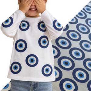Könnte beinhalten: Ein weißes Langarmshirt mit einem durchgehenden Muster aus dunkelblauen, hellblauen und schwarzen kreisförmigen Nazar-Auge-Motiven auf der Vorderseite und den Ärmeln. Das Shirt wird mit einer blauen Jeans kombiniert und zeigt einen grafischen Druck.