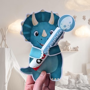 May include: A blue and teal dinosaur-shaped card with the text "bist eingeladen" on a white and red object. The dinosaur has a friendly face and is holding a round object with a white egg design.