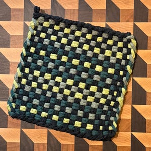 May include: A square potholder featuring a woven checkerboard pattern in shades of black, dark teal, sage green, and pale yellow. The edges are finished with a thick, braided border in matching colors, and a small loop is attached to one corner.