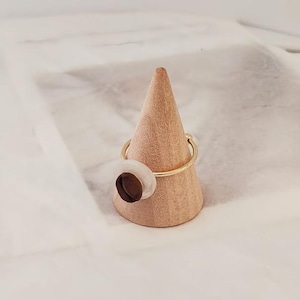 May include: A gold-colored ring with a circular design featuring a white and brown accent. The ring is displayed on a wooden cone-shaped stand against a white background. The ring's design is modern and minimalist.