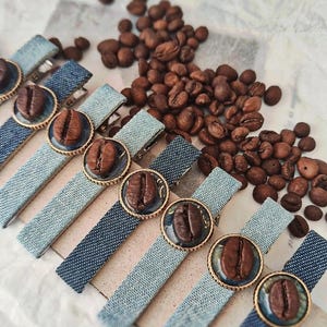 Peut inclure: Barrettes en jean avec un motif de grain de café. Chaque barrette a une bordure dorée autour du grain de café. Des grains de café sont éparpillés autour des barrettes.