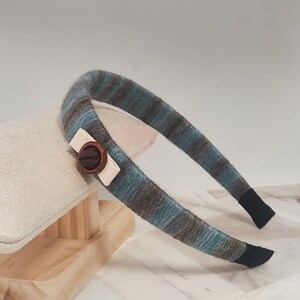 May include: A striped headband in shades of blue, gray, and brown. The headband features a decorative wooden button and a small white accent. The ends of the headband are finished with black fabric.