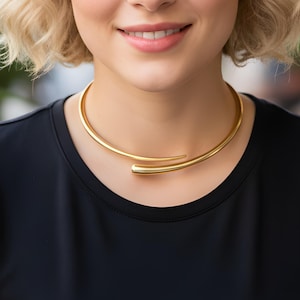 Può includere: Una collana a girocollo color oro dal design minimalista. La collana si curva attorno al collo e presenta un design elegante e aperto. Il gioiello è indossato sopra un top nero.