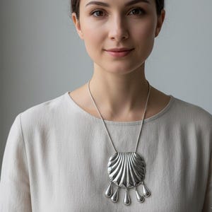 May include: A silver-toned pendant necklace featuring a large, stylized seashell design with a series of teardrop-shaped dangles. The necklace is worn on a silver chain. The woman in the image is wearing a light-colored top.