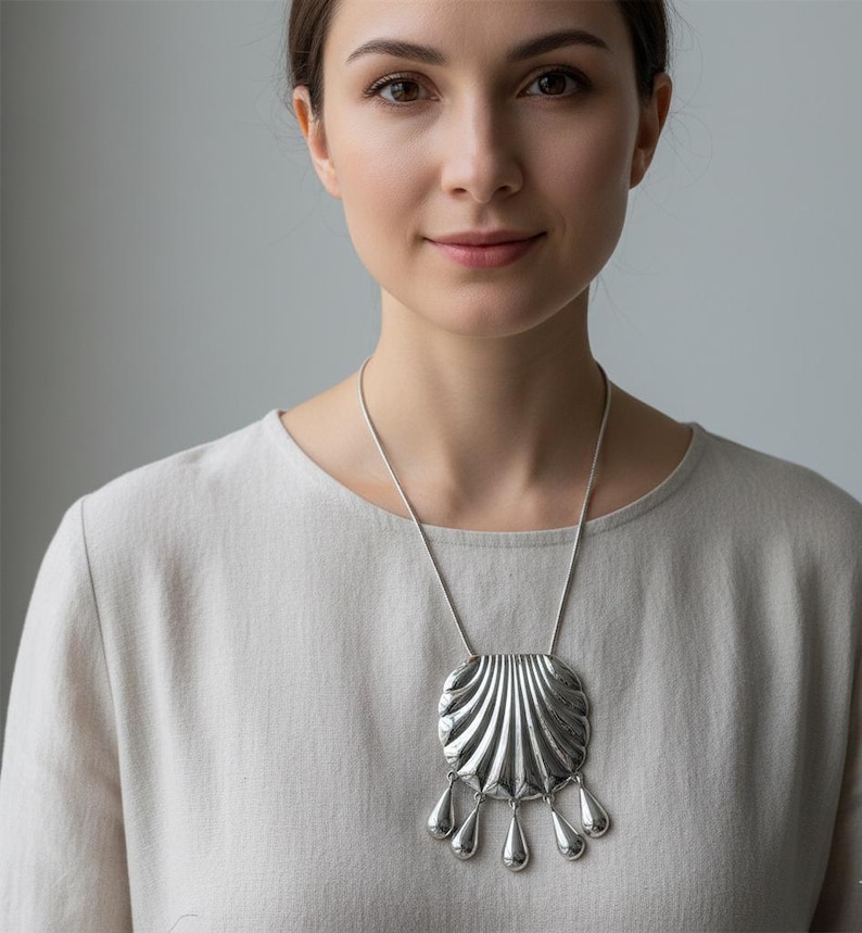 May include: A silver-toned pendant necklace featuring a large, stylized seashell design with a series of teardrop-shaped dangles. The necklace is worn on a silver chain. The woman in the image is wearing a light-colored top.