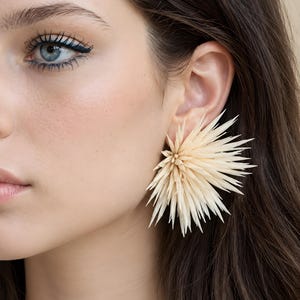 May include: A large, cream-colored statement earring featuring a dense, radial arrangement of thin, pointed, quill-like structures that fan out from a central point, creating a textured, spiky, and voluminous circular silhouette.