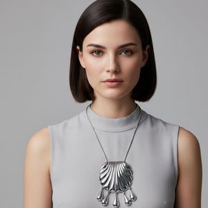 May include: A silver-toned pendant necklace featuring a square, fan-shaped design with five teardrop dangles. The necklace is worn on a silver chain. The woman in the image is wearing a gray sleeveless top.