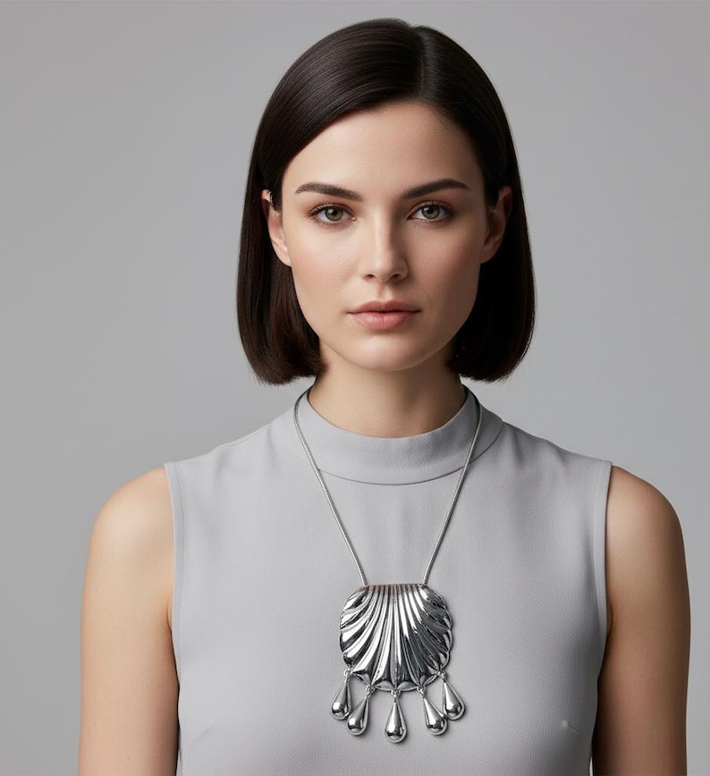 May include: A silver-toned pendant necklace featuring a square, fan-shaped design with five teardrop dangles. The necklace is worn on a silver chain. The woman in the image is wearing a gray sleeveless top.