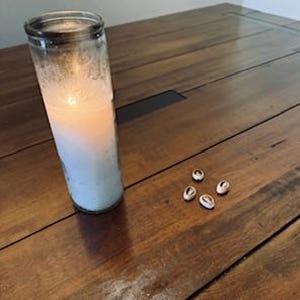 Op de afbeelding: Een brandende witte kaars in een heldere glazen pot en verschillende kleine, wit-bruine kaurischelpen liggen op een houten tafel. De kaars is cilindrisch en heeft een zachte gloed. De schelpen zijn verspreid in de buurt van de kaars.