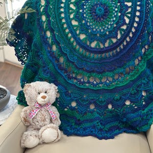 May include: A handmade, teal, blue, and green crocheted shawl with a circular design. The shawl is draped over a chair, next to a plush teddy bear with a pink and white ribbon. The crochet work features intricate patterns and openwork details.