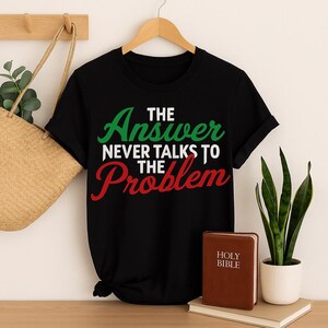 May include: Black t-shirt with the text "THE Answer NEVER TALKS TO THE Problem" in green, white, and red. A brown book with "HOLY BIBLE" printed on the cover is on a stack of books. A woven tote bag and a potted plant are also in the image.