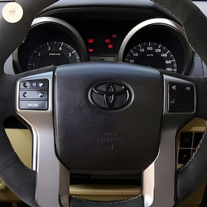Puede incluir: Primer plano de un volante negro Toyota con detalles plateados. El volante muestra indicadores, botones de control y el emblema de Toyota. El airbag está etiquetado. El volante es negro con costuras amarillas.