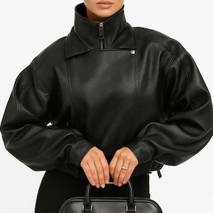 Peut inclure: Une veste noire texturée avec un col montant, une fermeture éclair frontale et un bouton-pression. La veste présente des manches longues et volumineuses avec des poignets ajustés et une coupe courte, offrant une allure structurée et grainée.