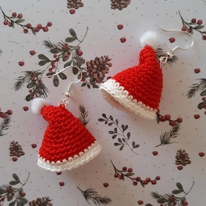 May include: A pair of festive crochet earrings shaped like miniature Santa hats. The hats are red with white trim and a small white pom-pom. The earrings hang from silver-colored fish hook ear wires. The background features a floral pattern.