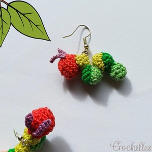 May include: Crocheted caterpillar earrings with red, yellow, and green segments. The earrings have a whimsical design with a hook closure. The earrings are handmade and feature a detailed, textured appearance. The earrings are a unique accessory.