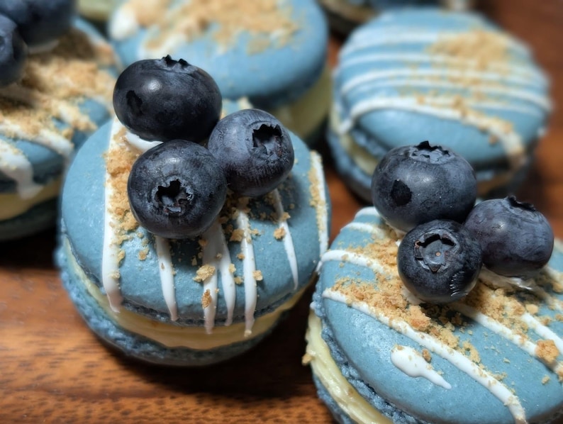 May include: Close-up of several light blue macarons, each topped with fresh blueberries and a sprinkle of crumbs. White icing drizzle decorates the tops. The macarons are arranged on a wooden surface, showcasing a dessert theme.