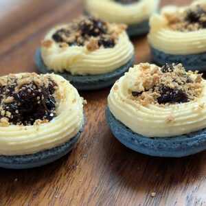 May include: Close-up of several macarons on a wooden surface. Each macaron has a blue shell, topped with a swirl of yellow cream, dark jam, and a sprinkle of crumbs. The macarons are arranged in a slightly blurred composition.