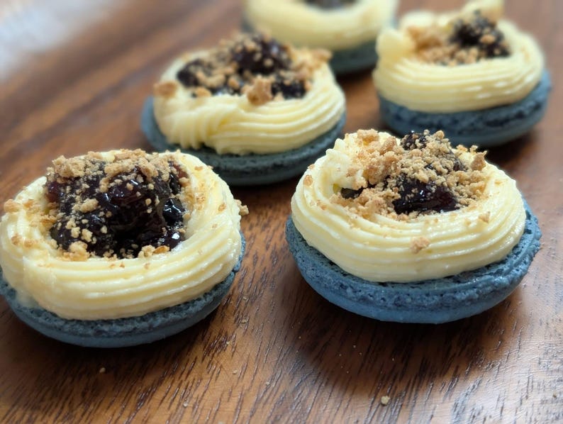 May include: Close-up of several macarons on a wooden surface. Each macaron has a blue shell, topped with a swirl of yellow cream, dark jam, and a sprinkle of crumbs. The macarons are arranged in a slightly blurred composition.