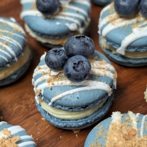 May include: Close-up of several blue macarons with white icing drizzle, topped with fresh blueberries and a sprinkle of crumbs. The macarons have a light yellow filling. The image showcases a dessert, likely homemade or from a bakery.
