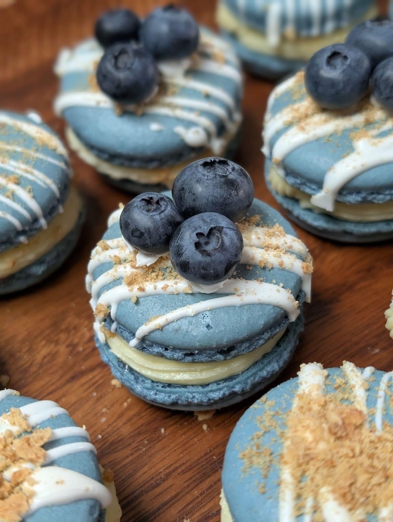 May include: Close-up of several blue macarons with white icing drizzle, topped with fresh blueberries and a sprinkle of crumbs. The macarons have a light yellow filling. The image showcases a dessert, likely homemade or from a bakery.
