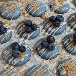 May include: Overhead shot of several blue macarons, each topped with fresh blueberries, white icing drizzle, and crushed cookie crumbs. The macarons are arranged on a light-colored surface, creating a visually appealing dessert display.