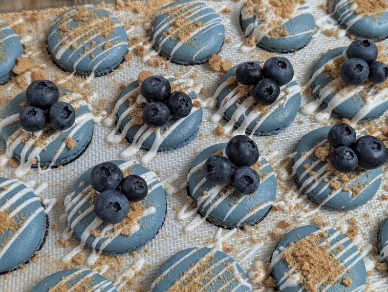 May include: Overhead shot of several blue macarons, each topped with fresh blueberries, white icing drizzle, and crushed cookie crumbs. The macarons are arranged on a light-colored surface, creating a visually appealing dessert display.