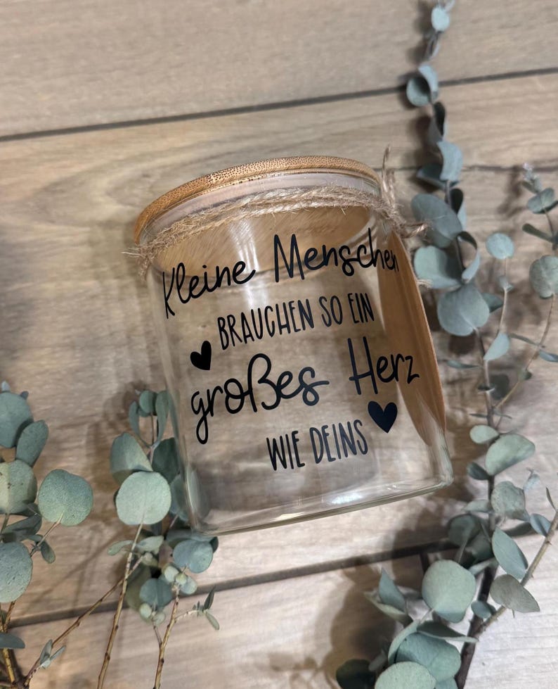 May include: A clear glass jar with a wooden lid and twine detail. The jar has black text that reads "Kleine Menschen brauchen so ein gro&szlig;es Herz wie deins" with heart symbols. Eucalyptus sprigs surround the jar.