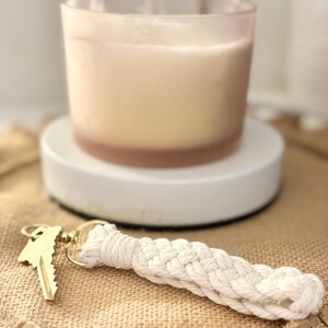 May include: A gold key with a woven white macrame keychain. A candle in a glass jar sits on a white circular base. The background is a tan burlap-like material.