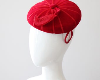 May include: A vibrant red hat with a round crown and a decorative feather. The hat features vertical ribbed detailing and a small loop on the side. The hat is displayed on a white mannequin head.