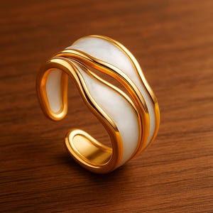 Peut inclure: Une bague dorée avec un insert en émail blanc. La bague présente un design ouvert unique avec des lignes fluides. Elle est posée sur une surface en bois brun chaud, soulignant sa forme élégante.