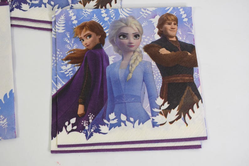 Pode incluir: Guardanapos de papel quadrados com personagens do filme de anima&ccedil;&atilde;o Frozen. Os guardanapos exibem Anna, Elsa e Kristoff contra um fundo floral azul e branco. Os guardanapos t&ecirc;m uma borda branca com uma faixa roxa.