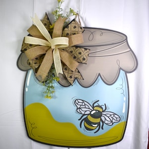 May include: A wooden honey jar-shaped sign with a bee illustration. The jar is light blue with a yellow base, a gray lid, and a decorative bow made of burlap and cream-colored ribbons. Greenery and yellow flowers are attached to the bow.